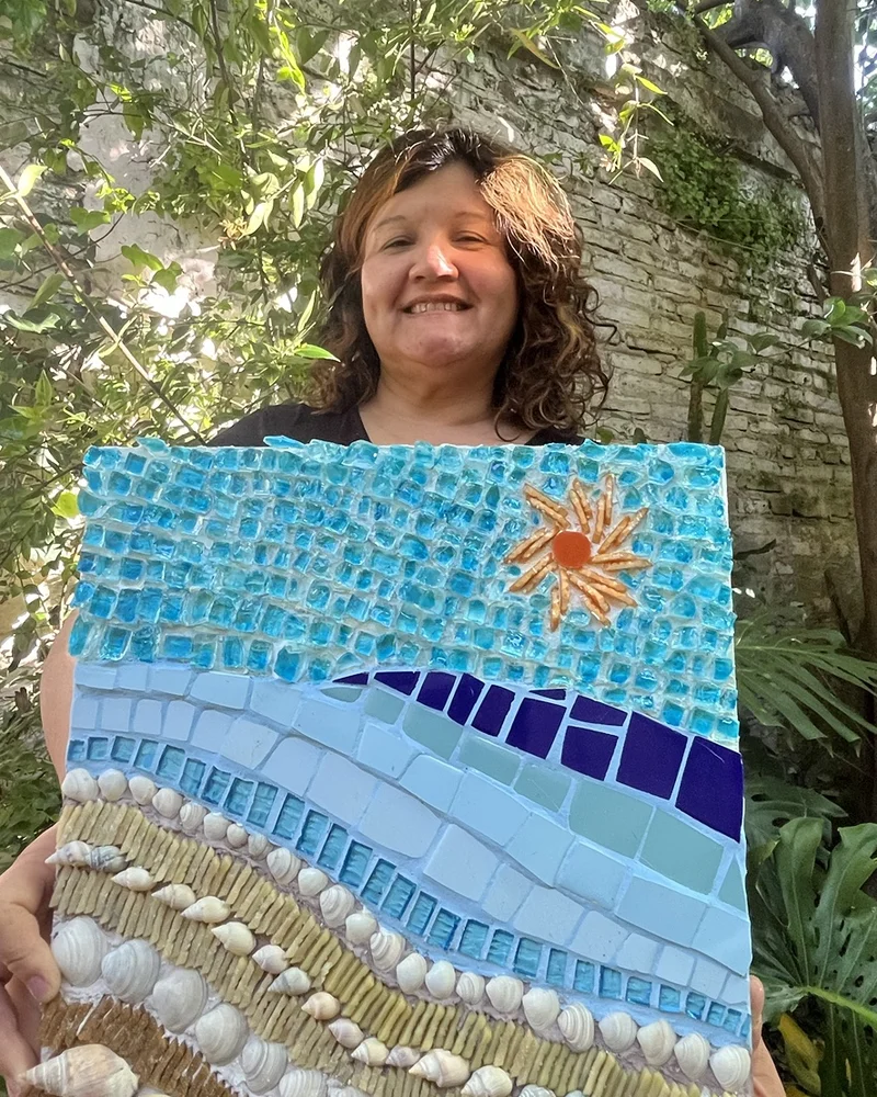María Alicia Molinas en un jardín, sosteniendo su mosaico de una orilla con caracoles